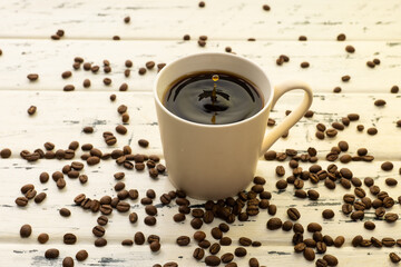 A mug of hot coffee with evaporation and steam and a small splash on a wooden table next to coffee beans