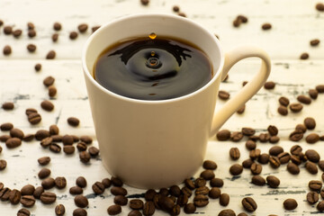 A mug of hot coffee with evaporation and steam and a small splash on a wooden table next to coffee beans