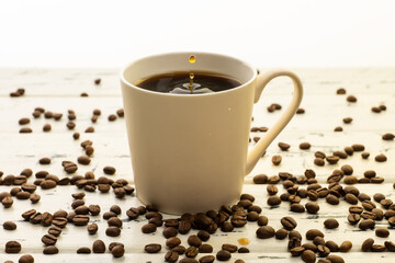 A mug of hot coffee with evaporation and steam and a small splash on a wooden table next to coffee beans
