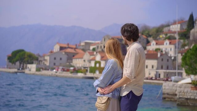A young couple of woman and man is hugging standing at the embankment of the city of Perast, a beautiful coastal town in the Boka-Kotorska bay. Travel to Montenegro concept. Slowmotion video