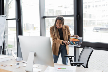 KYIV, UKRAINE - APRIL 27, 2022: african american businesswoman holding charts during video call near bitcoins on table.