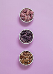 Three white bowl with colorful heirloom beans on a purple background. BLue, purple, brown scarlet runner beans. Top view. Copy space.