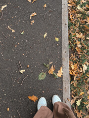 Women's legs in beige wide trousers and white sneakers stand on an asphalt path on which autumn leaves lie