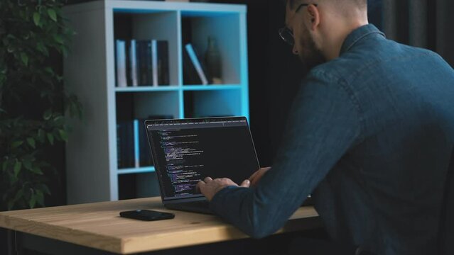 Bearded man in denim shirt writing code on laptop, young programmer coding soaftware in dark office