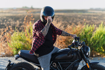 Chico joven tatuado con camisa a cuadros roja montando motocicleta por carretera solitaria al...