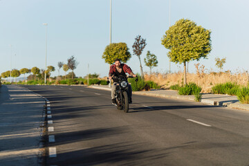 Chico joven tatuado con camisa a cuadros roja montando motocicleta por carretera solitaria al atardecer