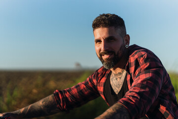 Chico joven tatuado con camisa a cuadros roja montando motocicleta por carretera solitaria al atardecer