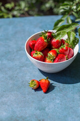 Bowl full of strawberries on blue background under sunlight. Concept of healthy foods and fruits.