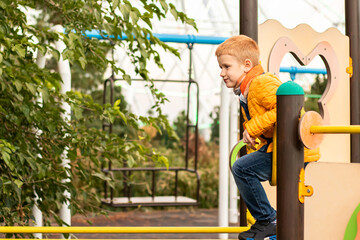 Red-haired cheerful teenager boy in yellow clothes having fun, playing, climbing on simulators on modern playground in city park