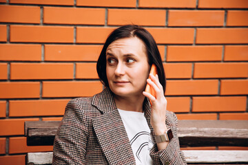 A girl in a brown jacket sits on a bench and talking on the phone. Portrait of a dark-haired girl in the park, front view