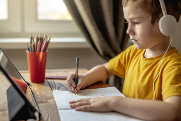 The child is a boy in a yellow T-shirt with headphones doing homework on a tablet. Concept of remote school education.