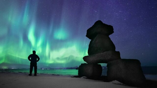 Timelapse of northern lights in Iceland - aurora borealis with man silhouette. Man in winter and ocean mountains shore with white snow - time lapse 4K. Unique weather green night starry sky