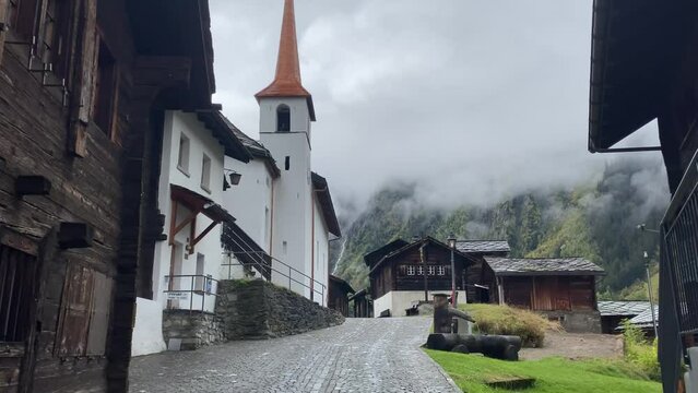 Old Village Of Blatten Bei Naters And Church With Mountains Shrouded By Clouds In The Background In Switzerland. - wide