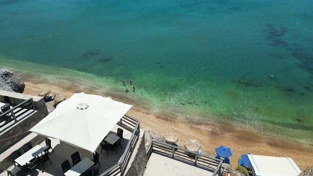 Tilt down drone view: restaurant terrace overlooking a sandy beach with parasols on a Greek island