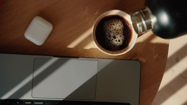 Top Down Shot Of Hot Coffee Getting Poured From Thermos In A Mug Next To Laptop In Home Office. 4K. Morning Sun. Cinematic