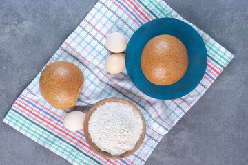 Flour bowl, eggs and buns on a towel on marble background