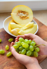 a woman's hand with green grapes, plate of melon slices on a wooden background, concept of fresh fruits and healthy food