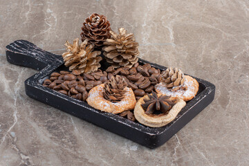 Pile of cookies, coffee beans, anise, and pine cones on a black tray on marble background