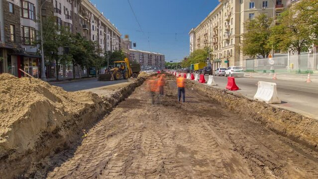 Workers in orange uniform digging and moving soil time lapse on road construction site. Industrial yellow excavator on a background. View of reconstruction of tram tracks on intersection