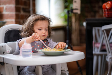 portrait caucasian baby girl about 2 years old in bib eating pasta from plate sitting high chair,...