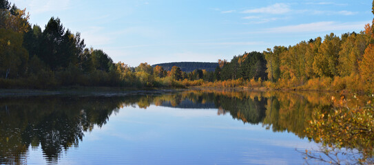 Autumn panoramic landscape. The two banks of the river are wooded. One side is lit by the sun. The second coast is in the shade. At sunset, the trees are reflected in the smooth surface of the water. 