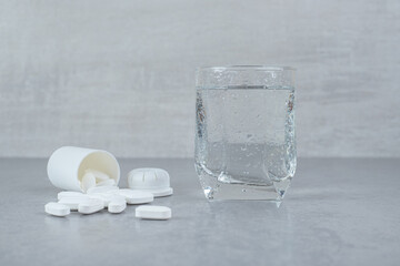 A small white jar of white pills with glass of pure water on gray background