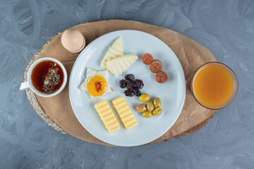 Breakfast with a cup of tea, a glass of juice, and a platter of cheese, egg, butter and sausage slices on a wooden board on marble background
