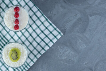 Bowls of milky rice garnished with raspberries and kiwi slice on a tablecloth on marble background