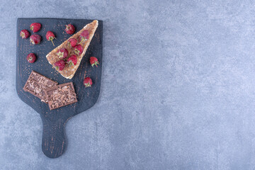 Black wooden board with chocolate plates and raspberry decorated cake slice on top on marble background