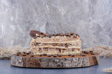 Appetizing slice of cake with coffee beans and chocolate bits, topped with a biscuit on a wooden board on marble background