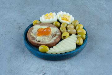 Breakfast platter containing boiled eggs, cheese slice, butterbrot and green olives on marble background