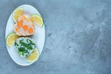 Portions of cooked rice with greens and carrots, garnished with lemon slices on a platter on marble background