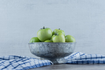 Old bowl filled with green tomatoes on a towel on marble background