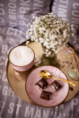 Cup of coffee with foam, pink saucer with pieces of chocolate on the golden tray on the grey bed, glasses and white flowers