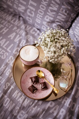 Fototapeta na wymiar Cup of coffee with foam, pink saucer with pieces of chocolate on the golden tray on the grey bed, glasses and white flowers