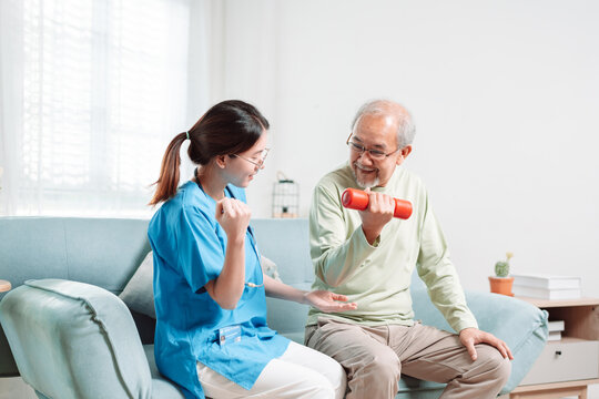 Asian Senior Old Man Doing Physiotherapist With Support From Caregiver. Mature Male Sitting On Sofa In Living Room Using Dumbbell Workout Exercise. Healthcare Medical Concept.