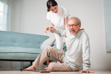 Fototapeta premium Asian woman daughter or granddaughter helping senior male from falling on the ground in living room at home. Elderly old mature grandfather having an accident heart attack after doing physical therapy