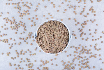 Brown lentil filled into a small bucket sitting in the midst of scattered grains on marble background