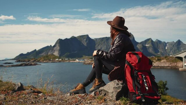 Girl with a big red tourist backpack sits on a rock overlooking the sea and enjoys a magnificent view lofoten mountains