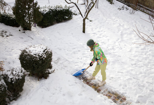 unrecognizable young woman in bright warm sportswear clears the path in the yard from snow after a heavy snowstorm using a large side shovel. Winter season, bad weather, cold, snow. Top view