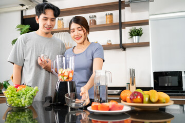 Happy Asian couple enjoy prepare cut fruits and vegetables ingredients in blender machine for making healthy vegan smoothie on the kitchen counter. Couple making vegan smoothie together at home.