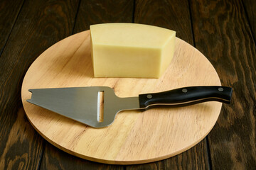 Cheese knife and a piece of hard cheese on a round wooden cutting board in close-up, shallow depth of field
