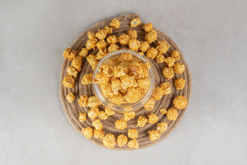 Glass bowl placed on a knitted trivet, filled and encircled with caramel coated popcorn on marble background