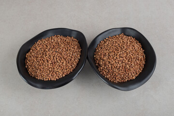 Two black bowls filled with raw buckwheat on marble background