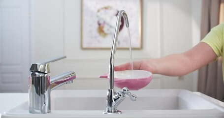 Child’s hands in the kitchen draw filtered water into a pink cup. Water flows from a small filter faucet. The filter faucet is in the center, medium shot.4K