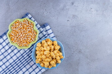 A bowl of caramel popcorn next to a bowl of corn grains on a towel on marble background