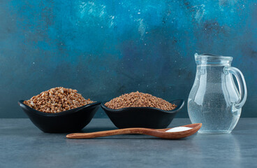 Cooking ingredients of two bowls of buckwheat, a small jug of water and a spoonful of salt on blue...