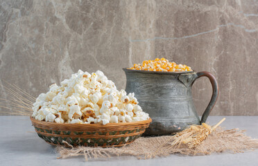 A jug of corn grains and a single wheat stalk next to a weaved basket of popcorn on a piece of...