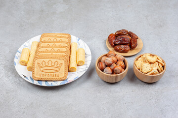 Bowls of dates, hazelnuts and crackers next to biscuits lined up on a plate on marble background
