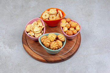Four colorful bowls of crispy biscuits and cookie chips on wooden board on marble background